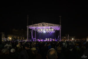 Crowd at a nighttime outdoor concert facing a brightly lit stage labeled The Stone Pony Summer Stage with purple and white lights illuminating the performers.