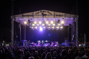 Nighttime outdoor concert with a brightly lit stage and a dense audience facing the performers on The Stone Pony Stage.
