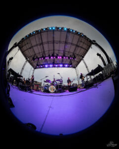 Live band performing on an outdoor stage under purple-blue lights, captured with a fisheye lens for a wide, circular view.