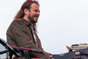 Man with long hair and beard playing a keyboard outdoors, wearing a brown jacket and smiling.