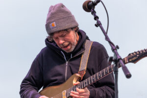Male guitarist wearing a gray knit beanie and dark hoodie, playing a gold electric guitar on stage with a microphone nearby.