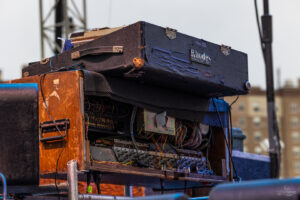 Worn Rhodes keyboard amplifier on a weathered wooden cabinet with tangled cables, outdoors on a stage.