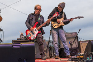 Two male musicians perform on an outdoor stage, one playing a red bass and the other a cream-colored electric guitar, with amps and cables nearby.