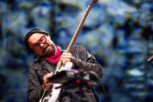 Musician in a black beanie and glasses leans into a white electric guitar on stage, with a blue abstract background.