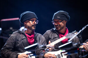 Two guitarists on stage wearing black beanies and red scarves, playing white electric guitars and smiling at each other.