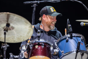 Smiling drummer with a green cap and beard behind a red drum kit with a large cymbal on the left.