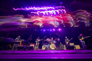 A live rock band on stage under purple lights, guitarists and a drummer playing with colorful wheel designs on the bass drums.