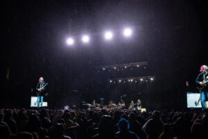Nighttime outdoor concert with a band performing on a brightly lit stage while a dense crowd watches from the foreground.
