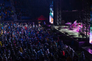 Large crowd at a nighttime outdoor concert, watching a band perform on a brightly lit stage to the right.