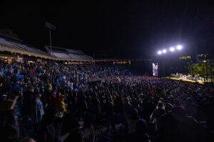 Nighttime stadium concert with a large crowd, performers on a brightly lit stage, and a tall video screen to the right.