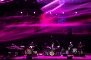 Live concert stage with a rock band performing under bright magenta and purple lights; keyboardist on the far left, a guitarist/singer center-left, drummer center, and two guitarists on the right, with amplifiers and colorful drum covers visible.