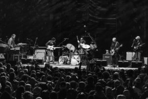 Black-and-white photo of a live band on stage performing before a dense crowd; center is a drummer with cymbals, flanked by guitarists and a keyboardist on the left, all amid amplifiers and microphones.