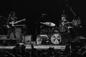 Black-and-white photo of a live band on stage: a guitarist left, a drummer right, with a cheering audience in the foreground.