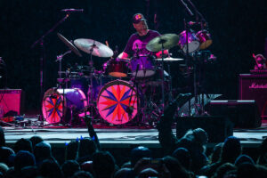 Drummer on a rain-soaked stage performing behind a colorful drum kit with starburst bass drums; cheering crowd in front.