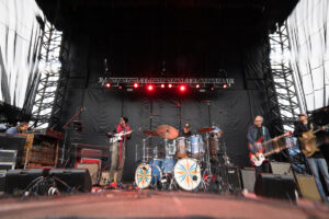 Band performing on an outdoor stage with a drummer center, guitarists on each side, and a keyboardist on the left under red stage lights.