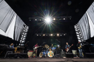 Band performing on an outdoor stage with drums in the center and guitarists on each side under bright overhead lights, colorful sunburst drum art visible on the toms and bass drums.