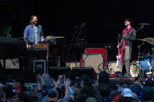 Two musicians perform on an outdoor stage: a keyboard player left and a guitarist right, with a cheering crowd in front.