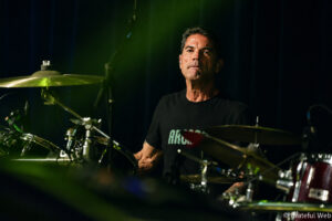 Drummer on stage behind a shiny drum kit with a focused expression under green stage lighting.