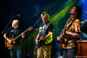Three guitarists performing on a dimly lit stage under blue and green lights during a live rock concert.