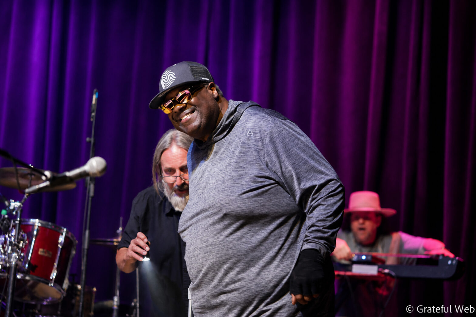 Male musician in a gray hoodie and cap performing on a stage with a purple curtain backdrop, sunglasses on, smiling as he leans forward.