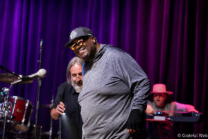 Male musician in a gray hoodie and cap performing on a stage with a purple curtain backdrop, sunglasses on, smiling as he leans forward.