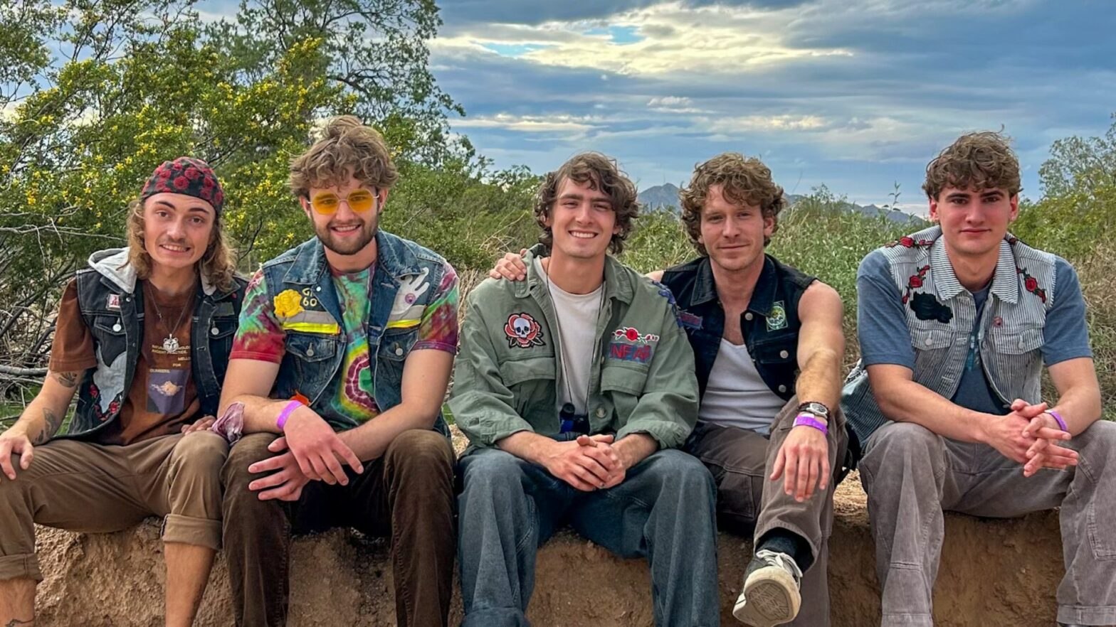 Five young men sit together on a dirt ledge outdoors, wearing patched denim jackets and casual clothing, smiling at the camera.