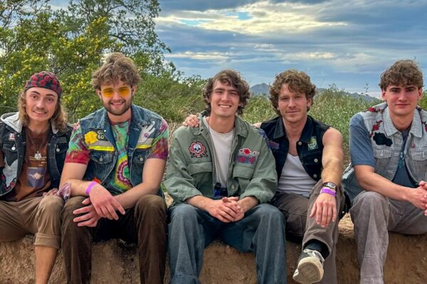 Five young men sit together on a dirt ledge outdoors, wearing patched denim jackets and casual clothing, smiling at the camera.