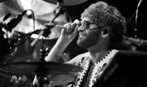 Bearded drummer in sunglasses holding a drumstick near his mouth while playing on stage in a black-and-white shot.