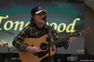 Male musician wearing a plaid shirt and cap, playing an acoustic guitar and singing into a microphone on stage.