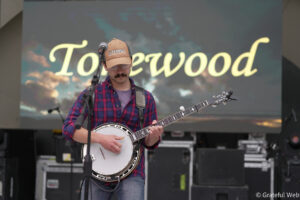 Male musician wearing a plaid shirt and brown cap plays a banjo on stage, microphone in front, with a large scripted word on the screen behind him.