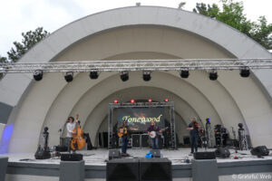 Musicians perform on an outdoor stage with arched white structures; a guitarist, bassist, and fiddler play as red stage lights glow, and a screen reads 'Tonewood'.