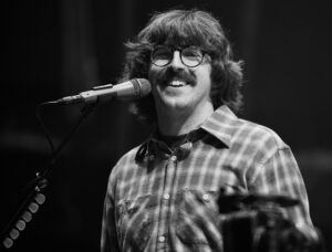 Male musician with round glasses singing into a microphone on stage, wearing a plaid shirt (black and white photo).