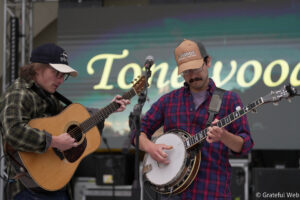Two male musicians perform on stage: one playing an acoustic guitar, the other a banjo, both wearing plaid shirts and baseball caps with logos.