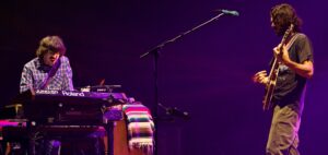 Two musicians performing on stage: a keyboardist in a plaid shirt on the left and a guitarist on the right under purple stage lighting.