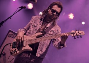 Musician with long hair and sunglasses playing a speckled bass guitar on a purple-lit stage, denim jacket visible.