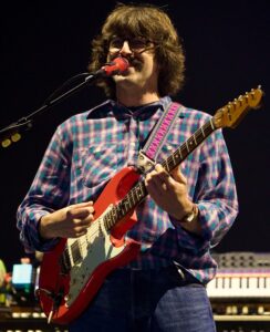Male guitarist wearing glasses and a purple plaid shirt playing a red electric guitar on stage.