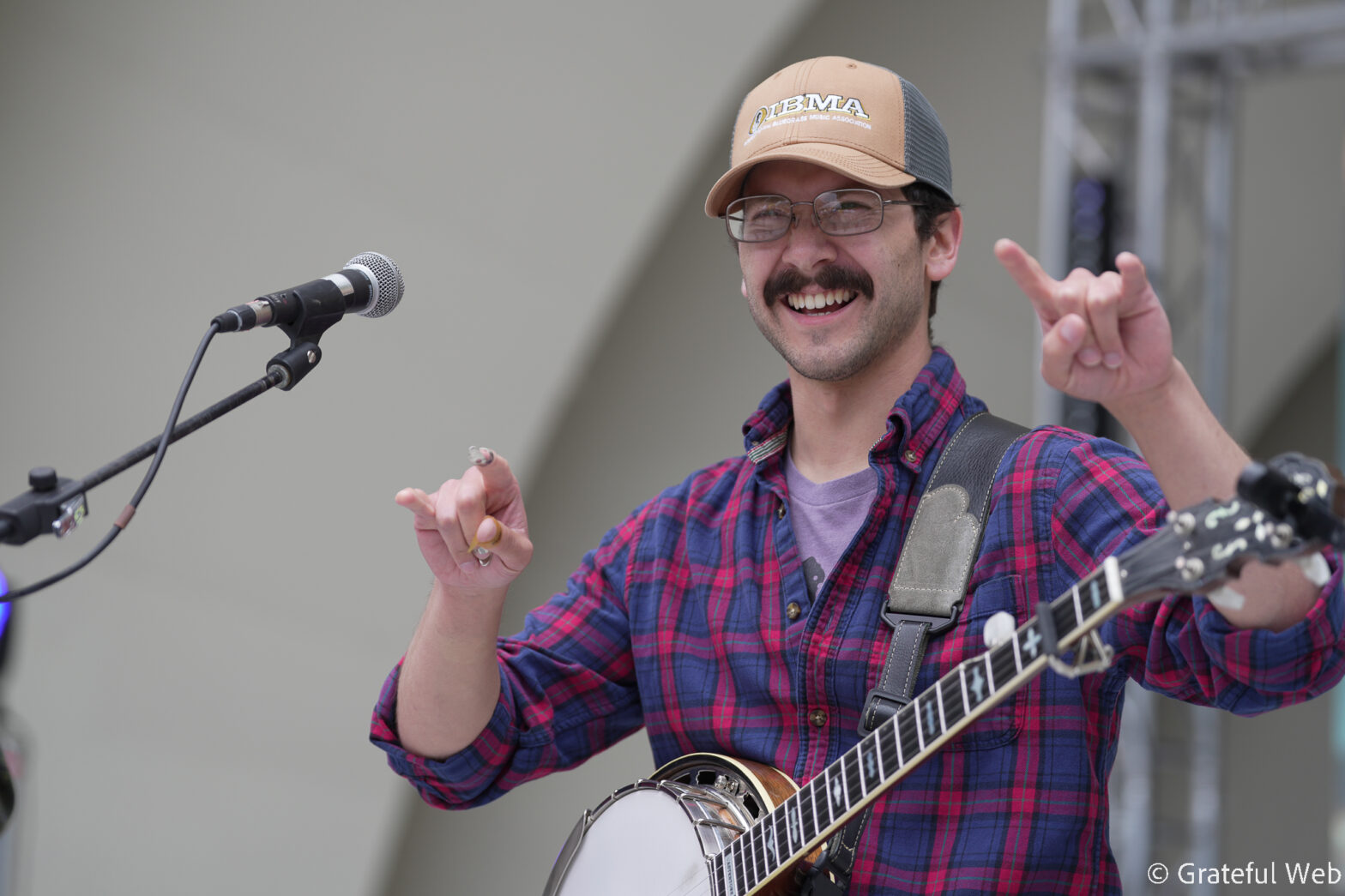Male musician singing into a microphone while playing an acoustic guitar on stage, wearing a tan cap and a red‑blue plaid shirt.