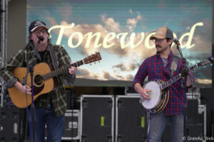 Two male musicians on stage: left guitarist sings into a mic while playing an acoustic guitar; right player holds a banjo and looks on.