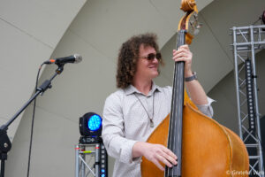 Musician with curly hair and sunglasses playing an upright double bass on a stage, microphone nearby and stage lighting in the background.