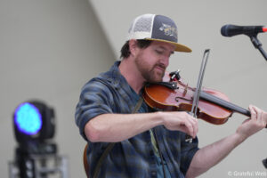 Man playing violin on a stage, wearing a gray mesh-back cap and blue plaid shirt, with a microphone nearby.