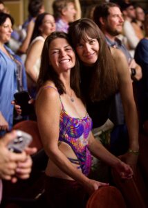 Two smiling women pose together in a crowded event, one in a colorful swimsuit, others blurred in the background.