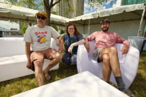 Three adults sit on white outdoor couches under a large event tent on a sunny day in the city park.