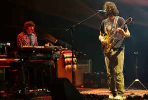 Two musicians perform on stage: a keyboardist on the left and a guitarist singing into a mic on the right.