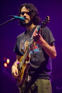 Male guitarist singing into microphone on stage, playing a sunburst electric guitar with purple stage lighting.