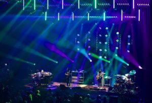 Concert stage with multiple musicians performing under vibrant blue, green, and purple light beams; keyboardist, guitarist, bassist, and drummer visible with audience in foreground.