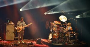 Live band on a dark stage: a bassist left, a drummer right, with bright spotlights and fog effects.
