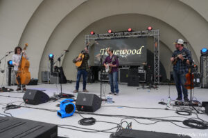 Four musicians perform on an outdoor stage with upright bass, guitar, banjo, and mandolin, backed by a large screen display.