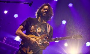 Male guitarist with long hair performing on stage, playing a sunburst electric guitar under purple stage lights.
