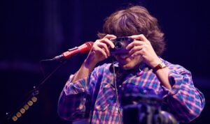 Male performer on stage holding a vintage camera to his eyes, wearing a purple plaid shirt with a red microphone beside him.