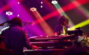 Two musicians on stage playing keyboards under bright magenta and purple lights.